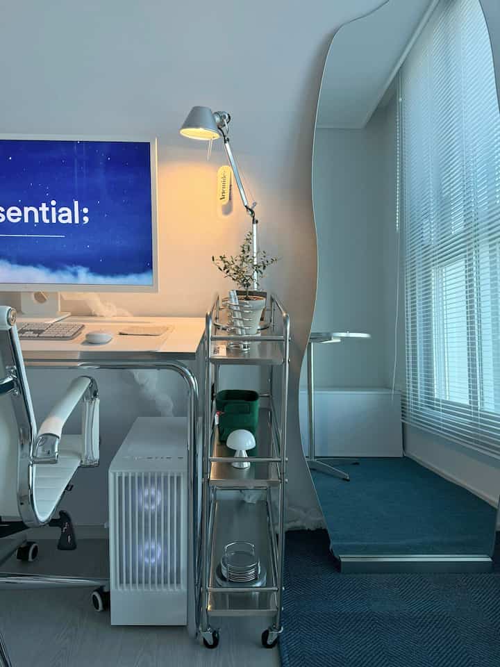 白と青のトーンのモダンな書斎、白いデスクとオフィスチェア、アルテミデの照明、青いカーペットと曲線的な全身鏡が配置された清潔感のあるインテリア