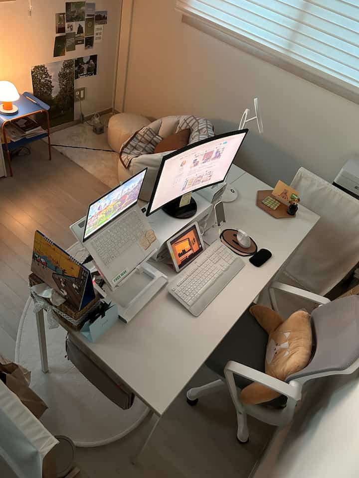 Bright white cozy home office workspace featuring a wide desk with monitors, laptop, and an office chair near a window