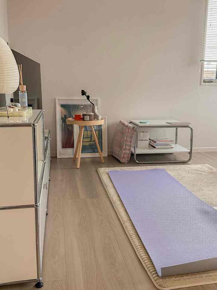 ベージュとナチュラルモダンのトーンが特徴のコンパクトな一人暮らし用リビング、ラベンダーのエクササイズマットとサイドテーブル、サイドボードが配置された清潔感のある空間