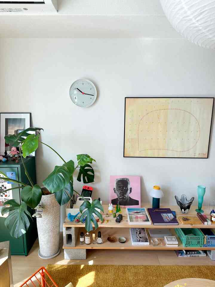 白い壁とナチュラルな木目調の棚があるリビング、モンステラの植物と時計、モダンなインテリアが配置された空間