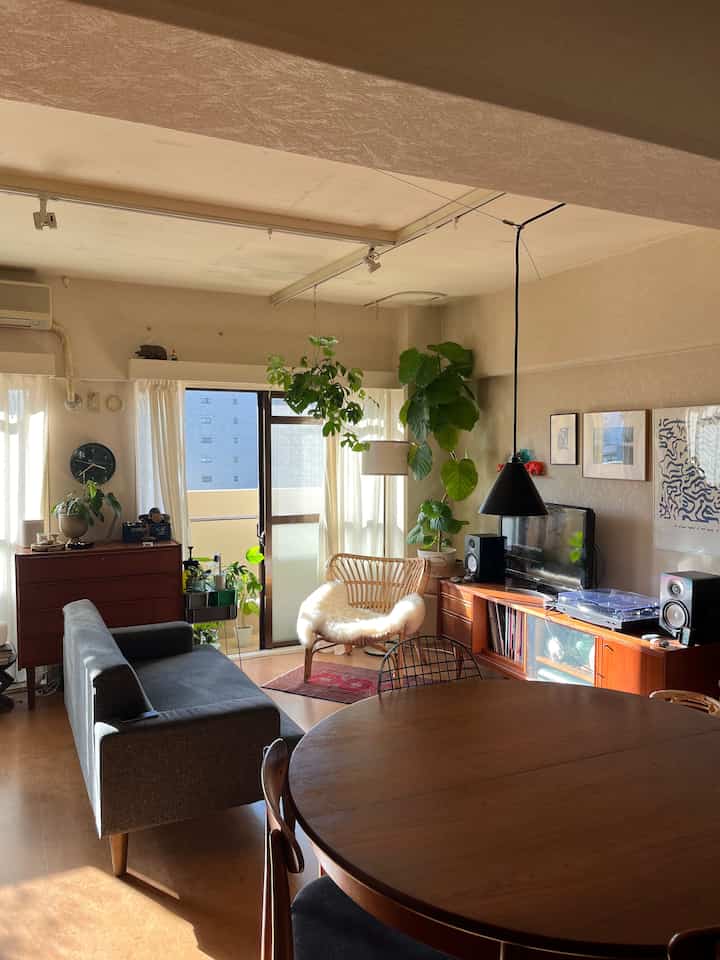 ライトブラウンの木目調とダークグレーのソファがある、植物と自然光が満ちた印象的なリビングダイニング空間