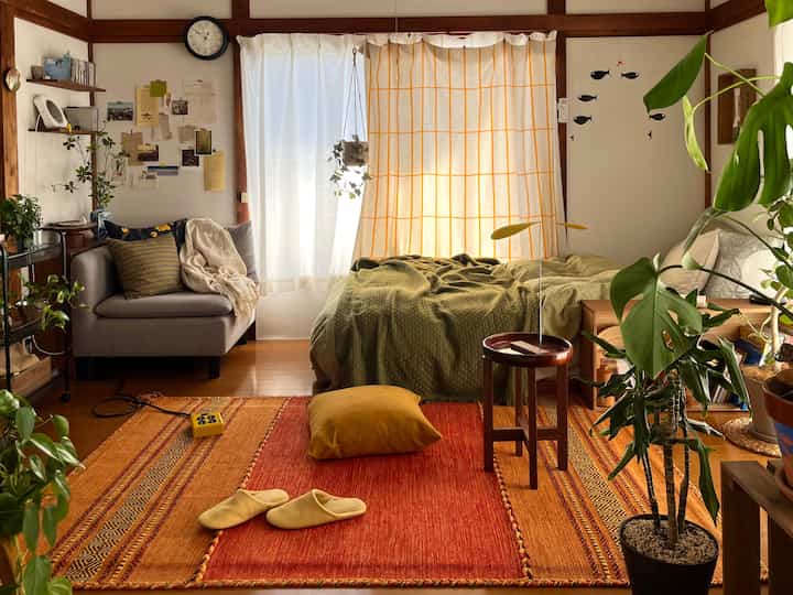 温かみのあるウッドトーンと豊かなグリーンの植物が調和した1人暮らしのワンルームリビング、オレンジのラグとグリーンのベッドリネンが印象的なナチュラルモダンな空間