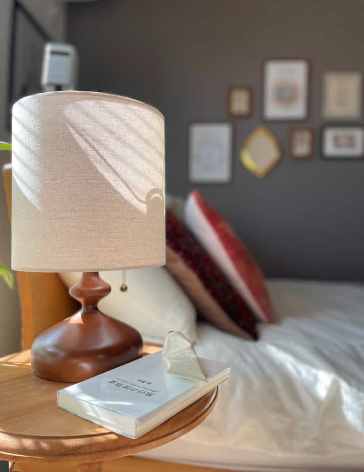 Natural wood tone and beige lighting blend in a cozy single-bedroom featuring a round bedside table with a table lamp and book.