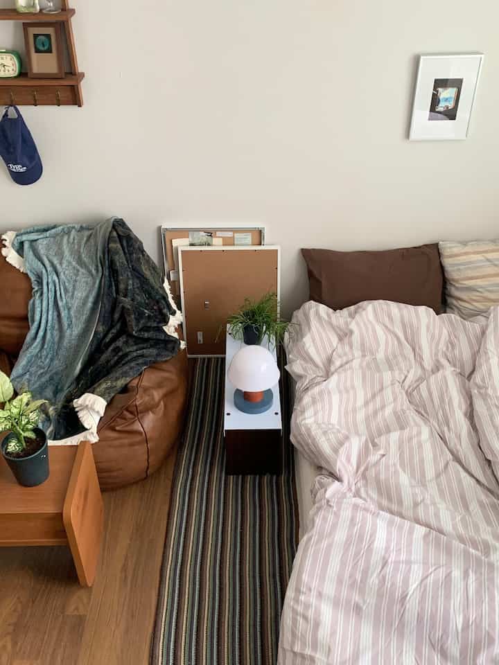 ブラウンの木目調床と白い壁のナチュラルでシンプルな寝室、茶色のビーンバッグとストライプの寝具、サイドテーブルに植物とテーブルランプが配置された空間