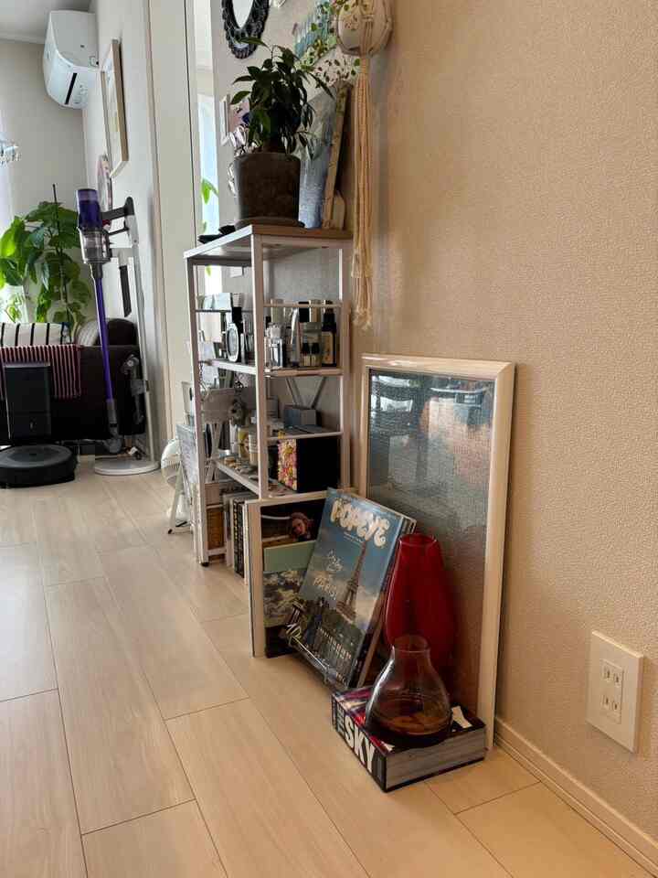 明るい木目調の床とベージュの壁を持つコンパクトなリビング、棚とソファ、赤い花瓶と植物が自然な雰囲気を演出しています