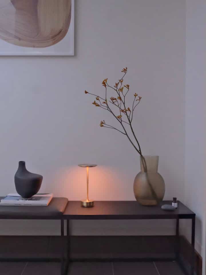 グレーの壁とダークブラウンのベンチがあるシンプルな玄関、テーブルランプと花瓶が調和した印象的な空間