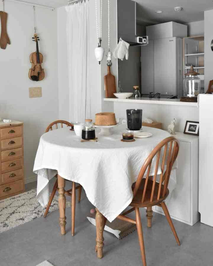 明るい白とナチュラルな木目調が調和した小さなワンルームのキッチンダイニング空間、木の椅子と白いリネンのテーブルクロスにコーヒーとケーキが置かれている雰囲気のあるインテリア