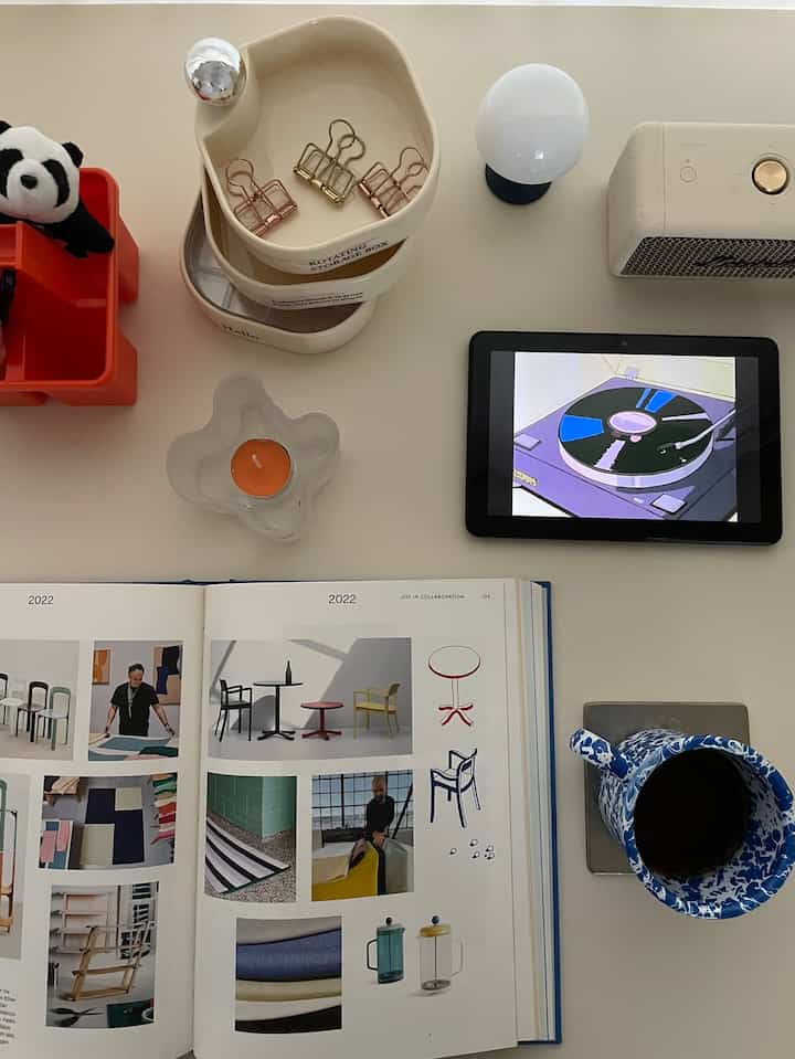 White-toned home office workspace featuring a table mat with an open book, tablet, and table lamp arranged neatly