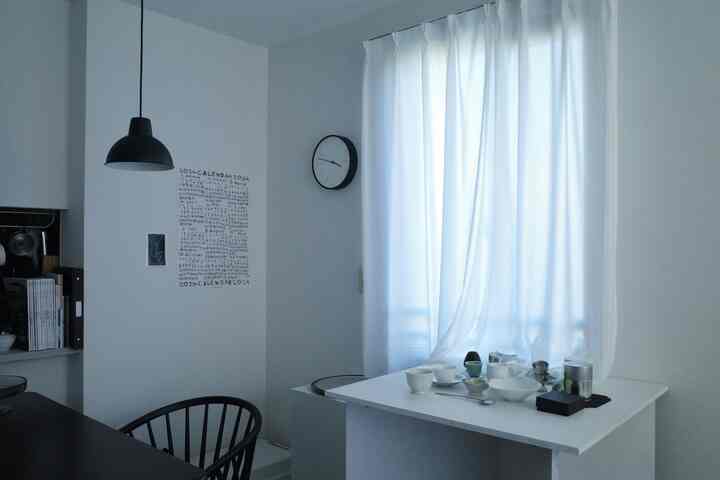 Bright white-toned dining room featuring a white dining table, black chairs, sheer curtains, and wall clock creating a simple atmosphere