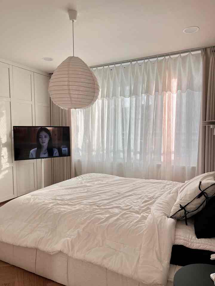 明るいホワイトトーンの寝室で、大きなベッドと透けるカーテン、モダンな照明が配された落ち着いた空間