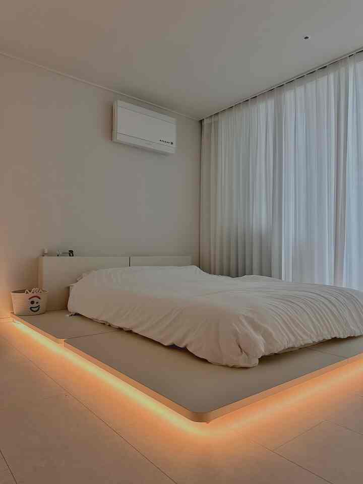 A white and beige toned bedroom featuring a low-profile DIY bed frame and indirect lighting creating a cozy atmosphere