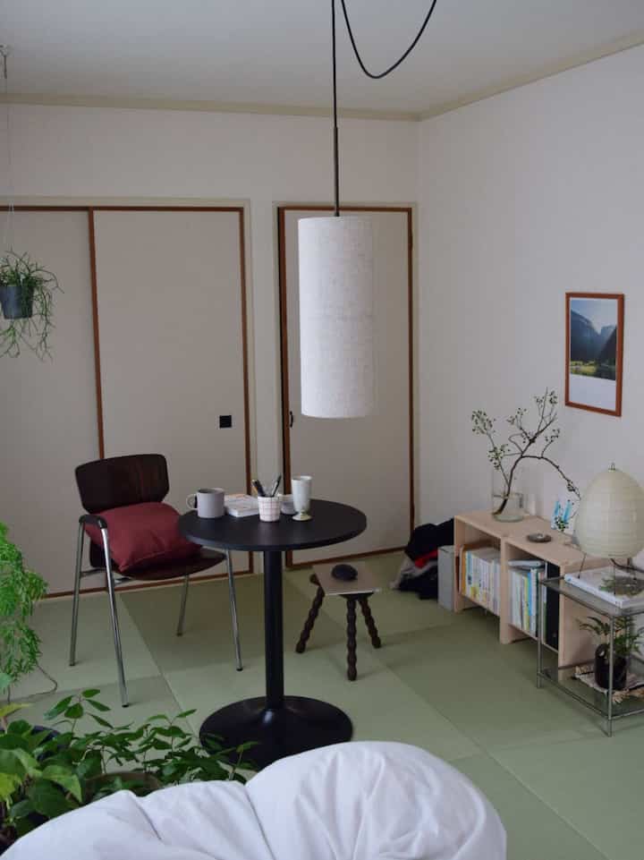 白い壁とナチュラルな木製棚が配置された狭い部屋、中央にはペンダントライトが吊るされた黒い丸いダイニングテーブルが置かれた北欧風のインテリア