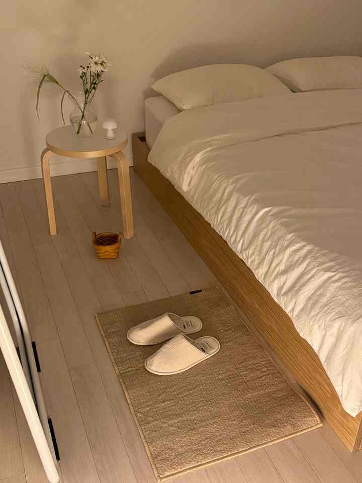 柔らかなベージュと木目調の寝室、木製スツールとスリッパが置かれた清潔で居心地の良い空間