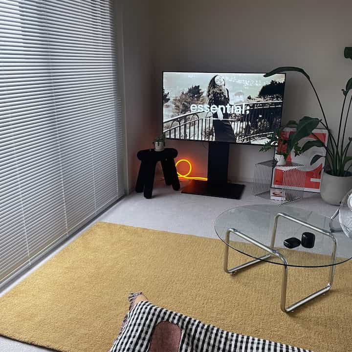 A modern living room in white and yellow tones featuring a large TV, transparent glass table, and yellow rug