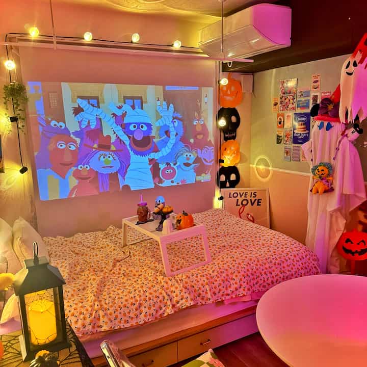 温かみのあるオレンジ色の照明が印象的な一人暮らしの寝室、プロジェクタースクリーンとハロウィンの飾りが配置されたホームシネマ風のインテリア