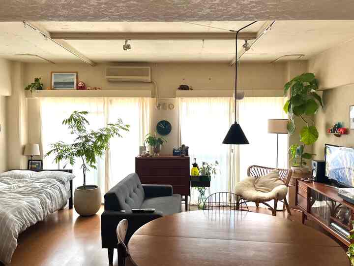 ベージュ調のワンルーム、ソファと木製のダイニングテーブル、多数の観葉植物が配置された雰囲気のある空間