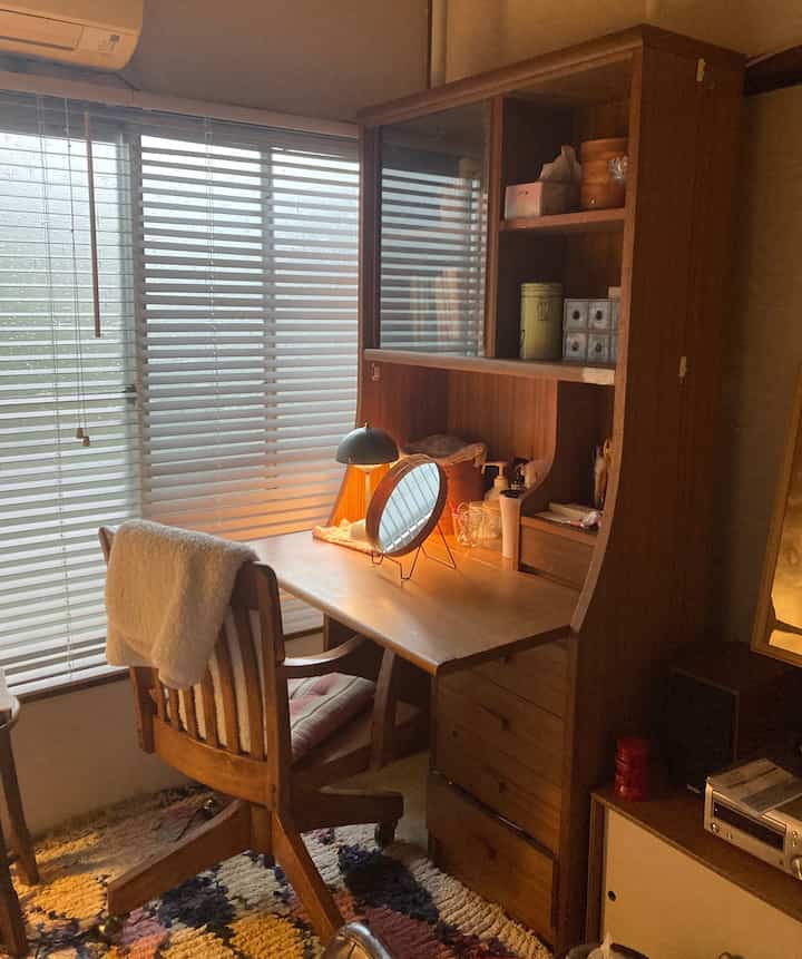 温かみのある木目調の狭い部屋に配置されたヴィンテージ風のドレッサーと椅子、窓には白いブラインドが掛かり落ち着いた雰囲気のインテリア