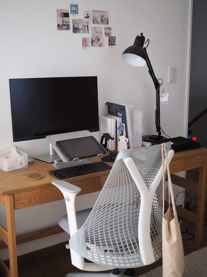 Simple and natural home office space with white walls and brown wooden desk, featuring Herman Miller office chair, monitor, and desk lamp