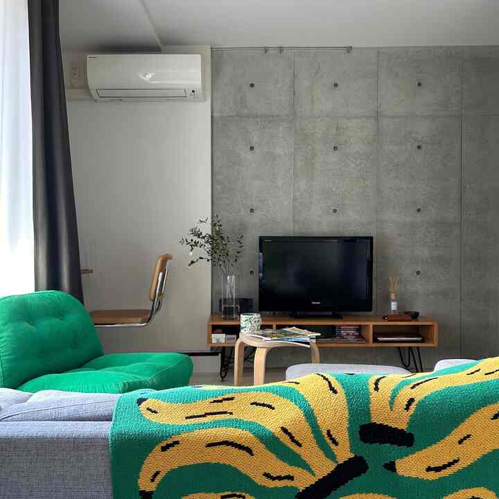グレーのコンクリート壁と木目調のテレビ台、緑のアームチェアとグレーのソファが配置されたミニマルなワンルームのリビング
