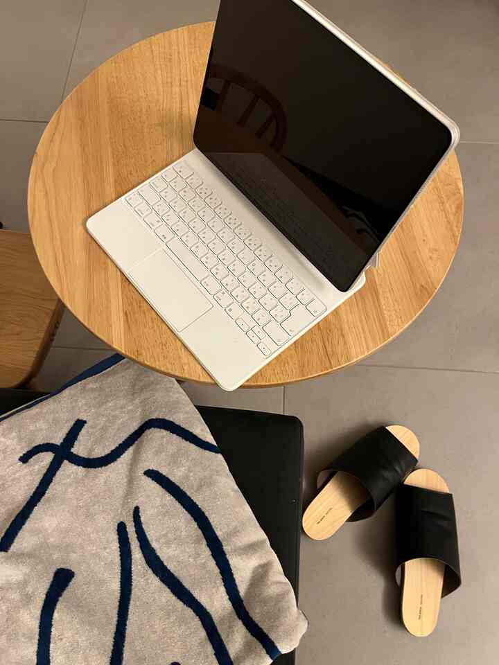 木目調の丸テーブルにiPad Proが置かれ、その隣に黒いスリッパとベージュ色のネイビーライン入りタオルが配置された、モダンでコンパクトなホームオフィス空間