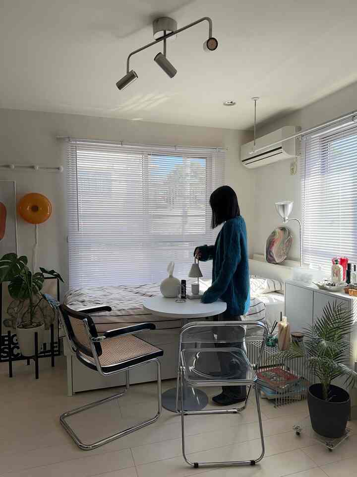 明るいナチュラル色の小さな寝室、ラタンチェアと折り畳みチェア、小さな丸テーブルが配置されたミニマルな空間