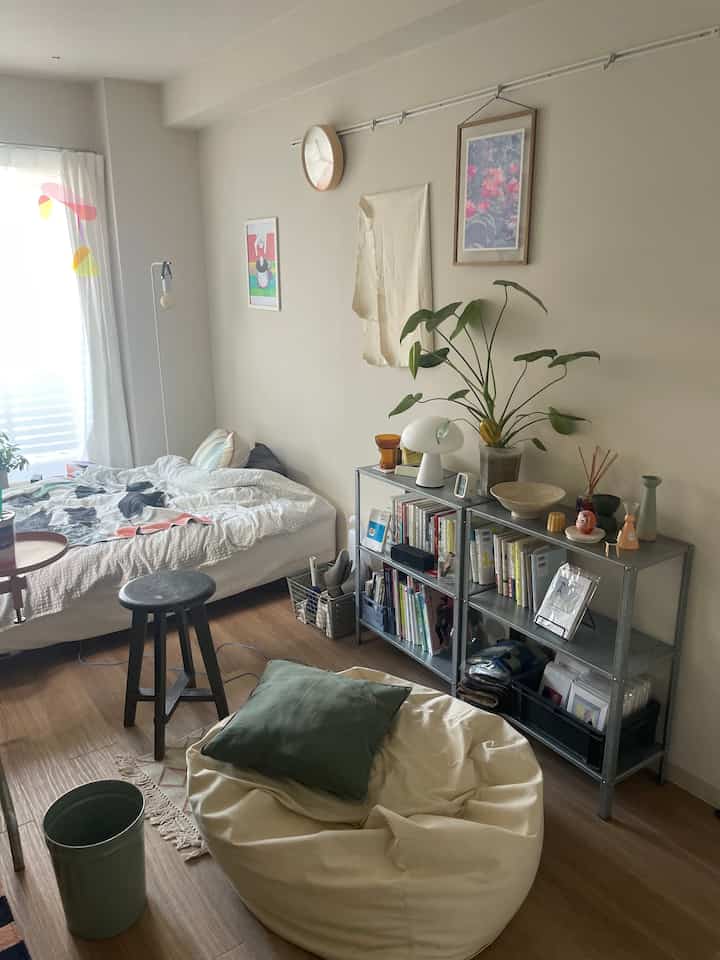 ベージュの色調の寝室、ベージュのビーズクッションと黒いスツール、ライト付きの金属製棚が配置された落ち着いた雰囲気のインテリア