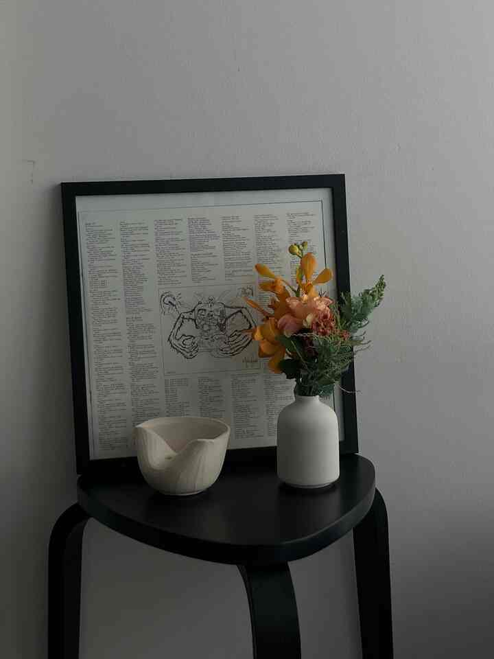 Simple natural style space featuring a black stool with a white vase of flowers and ceramic bowl against a white wall with a framed record lyric