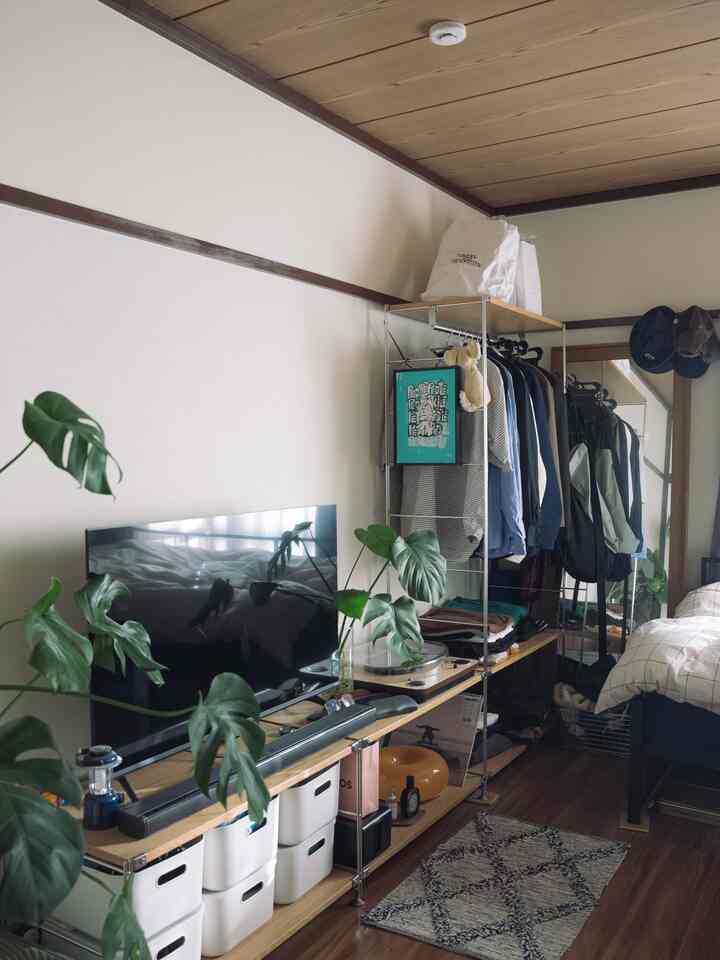 ナチュラルな木目調とブラックメタルの棚が調和するコンパクトな寝室空間、壁際に無印良品のユニットシェルフとテレビスタンドが配置されている