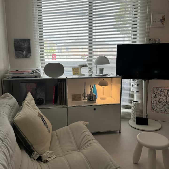 Bright white-toned living room featuring a minimal white sofa, TV stand, modular storage cabinet, and decorative accessories creating a simple atmosphere