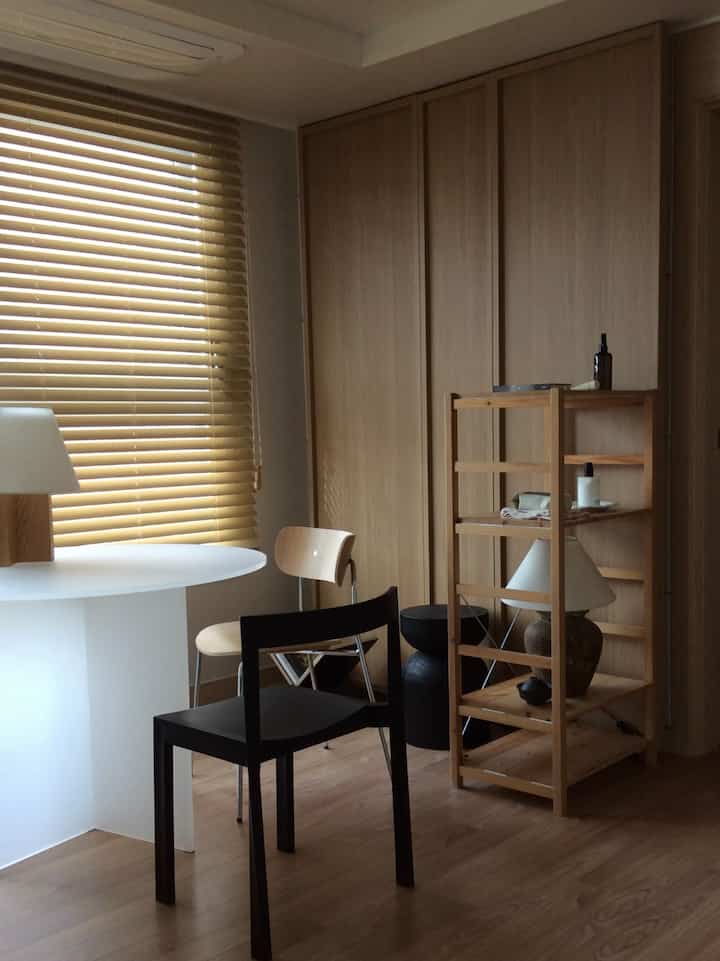 温かみのあるウッドトーンとモノトーンが調和する狭いリビング、ウッドパーティションと松の棚、ブラインドが配置された落ち着いたインテリア