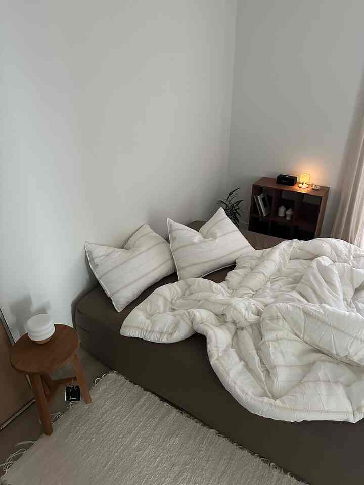 ホワイトの壁とブラウンのマットレスが調和した寝室、ウッドトーンの家具とホワイトの寝具が配置された雰囲気のある空間