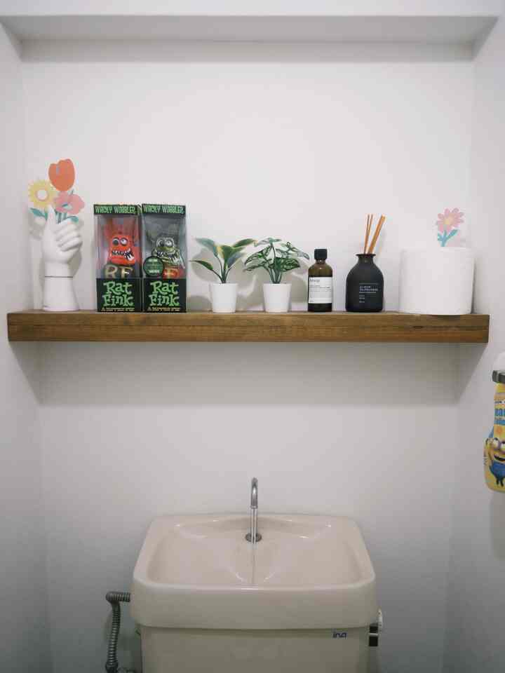 白を基調とした狭いトイレの壁にナチュラルな木製棚が設置され、小さな植物やディフューザー、トイレットペーパーが配置された落ち着いたインテリア