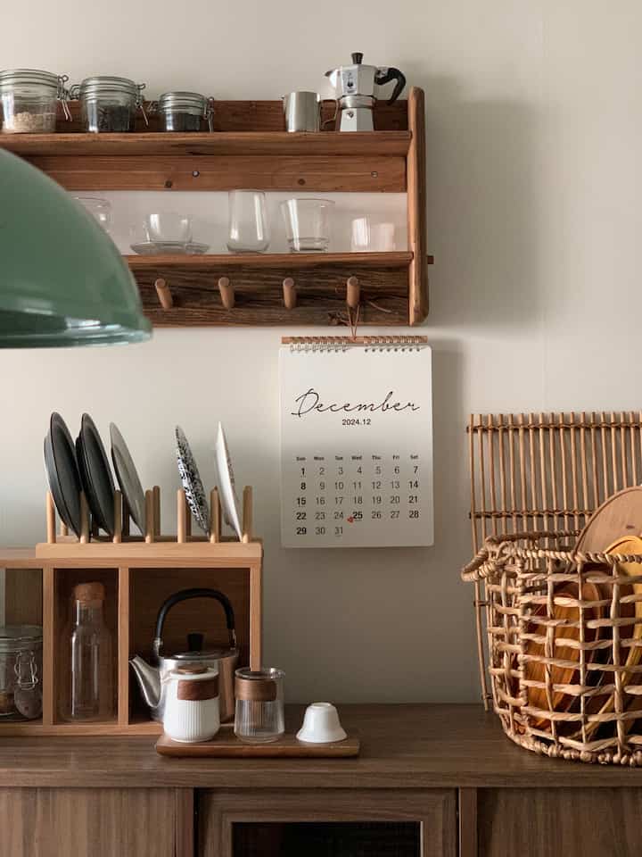 木目調とナチュラルカラーが調和したキッチンスペース、壁には2025年の壁掛けカレンダーが中央にかかり、木製棚と食器ラック、ラタンバスケットが配置された印象的な空間