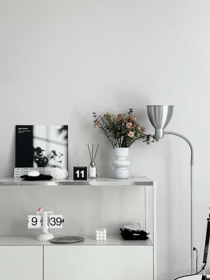 白と黒のトーンのワンルームインテリア、整頓された収納キャビネットとフリップクロック、花瓶、フロアランプが配置されたシンプルモダンな空間