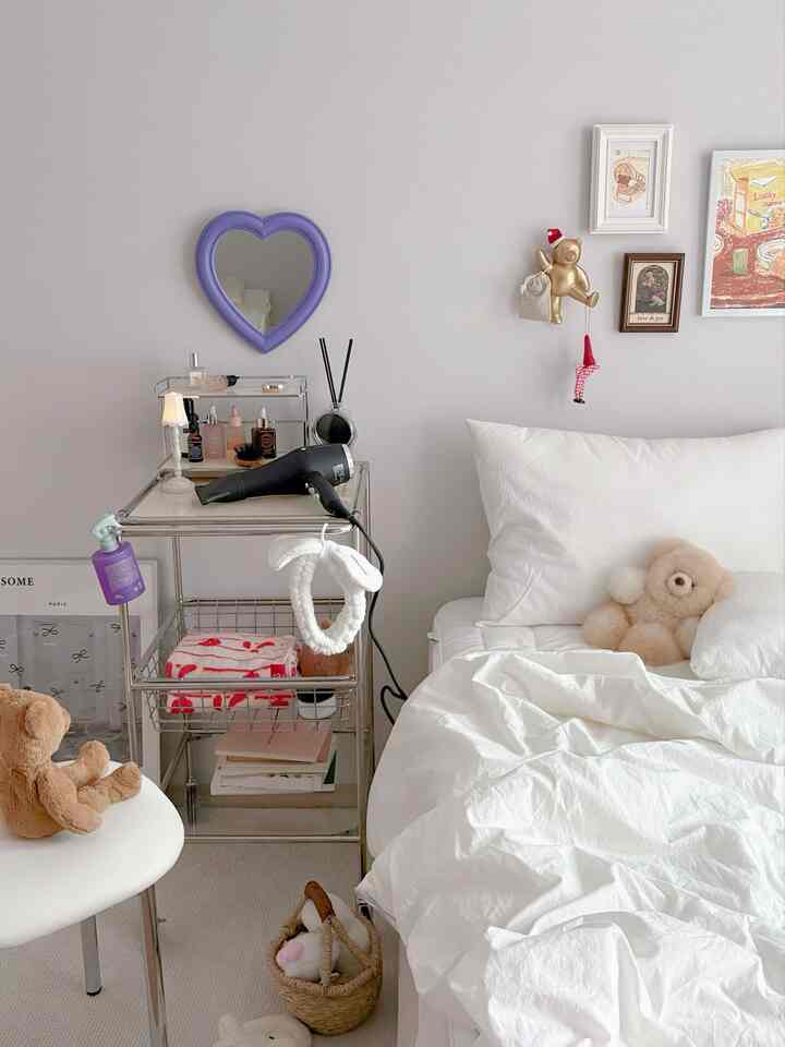White-toned bedroom featuring a simple metal nightstand, heart-shaped wall mirror, and cozy teddy bears
