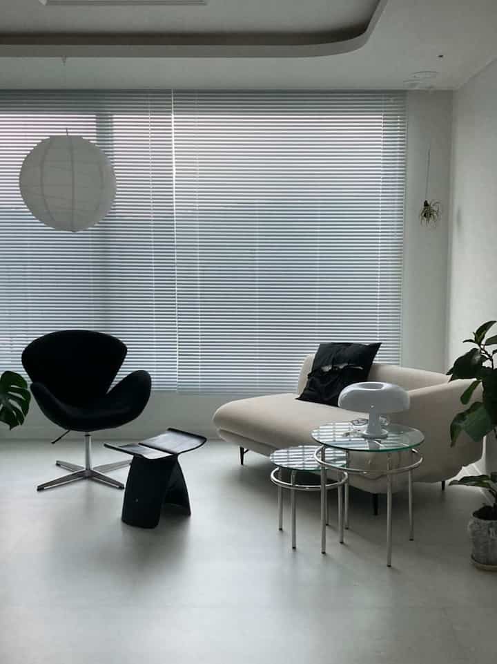 Bright white-toned living room featuring a beige sofa, black armchair, glass side tables, and plants in a simple modern setting