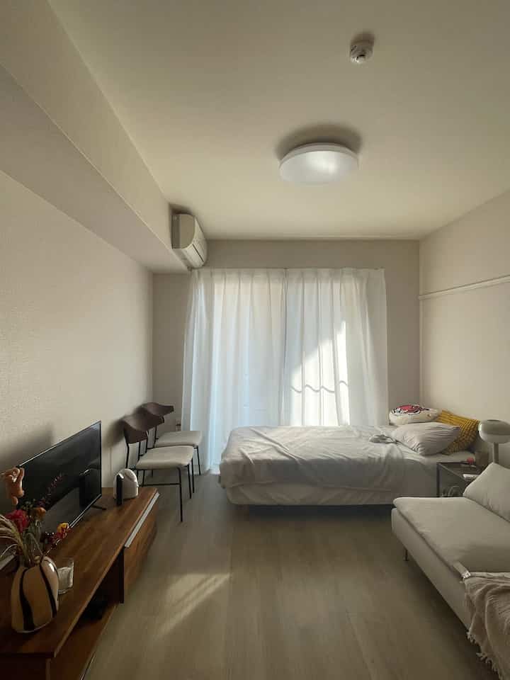 ホワイトとナチュラルな色調の寝室、シングルベッドとソファ、テレビ台が配置されたコンパクトなワンルームのインテリア