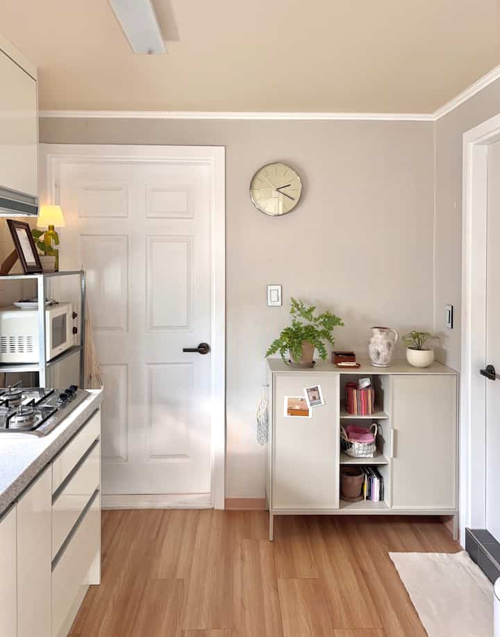 明るい木目調の床と白い壁が調和したコンパクトなキッチン、キャビネットと壁時計、観葉植物が配置された清潔感のあるミニマルなインテリア