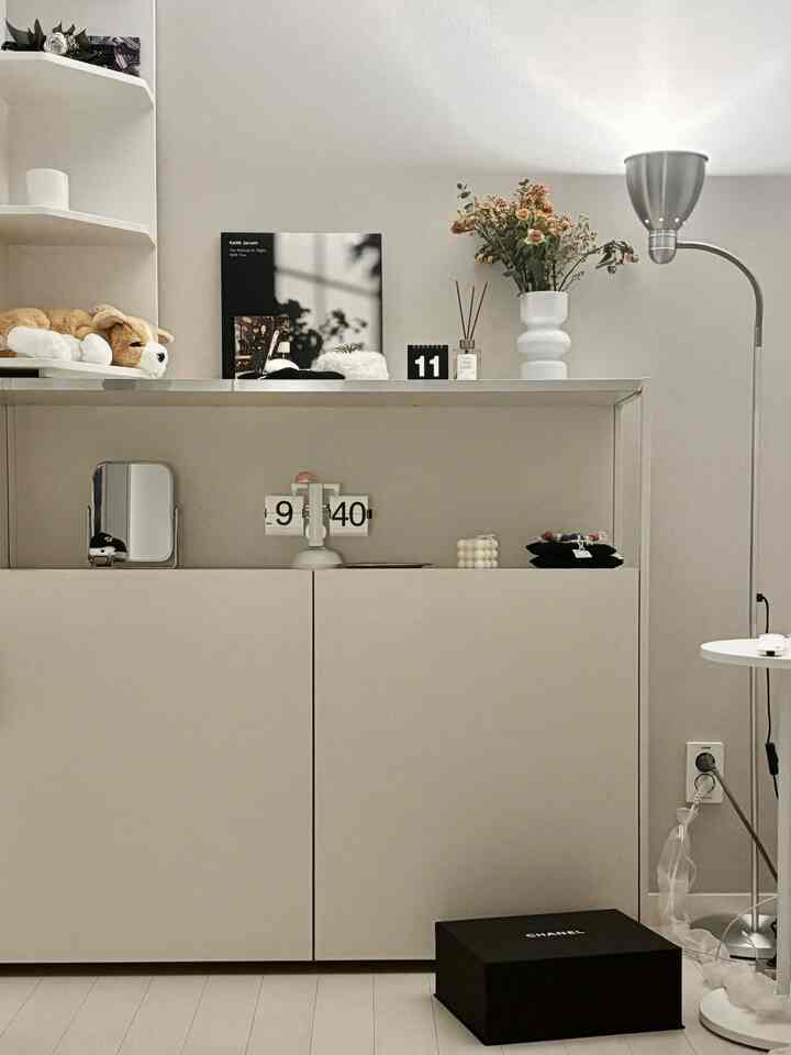Simple living room in white and beige tones featuring a large door cabinet, corner shelves, floor lamp, and various objets creating a cozy atmosphere