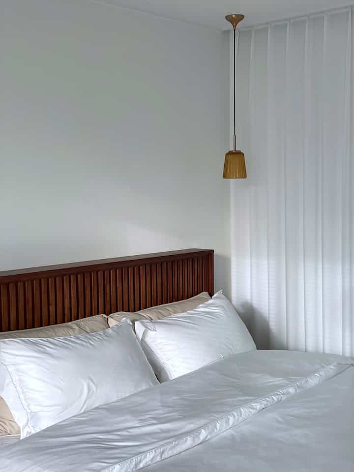 A neat hotel-like bedroom in white and beige tones featuring a wooden headboard bed and indirect pendant lighting