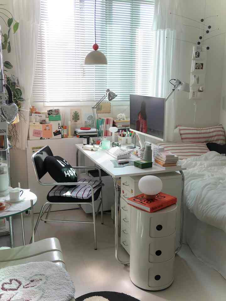 A compact white-toned bedroom featuring a desk with computer by the window, plants, and a bedside table, creating a cozy home office space
