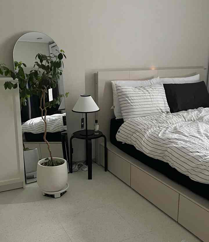 ホワイトとブラックのトーンの寝室、ストライプ寝具と収納付きベッド、スツールと植物が配置されたモダンなインテリア