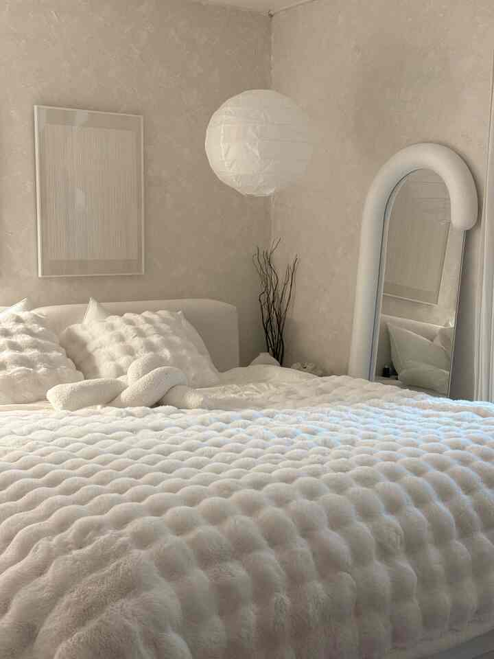 温かみのある白系の寝室、ふわふわのブランケットとクッション、アーチ型の鏡と紙製ペンダントライトが配置された雰囲気のあるインテリア