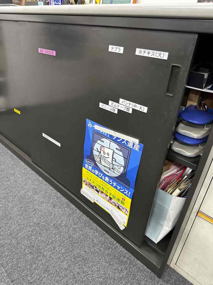 Black sliding office cabinet with multiple Japanese labels, adjacent blue storage containers, and a gray carpeted office floor area