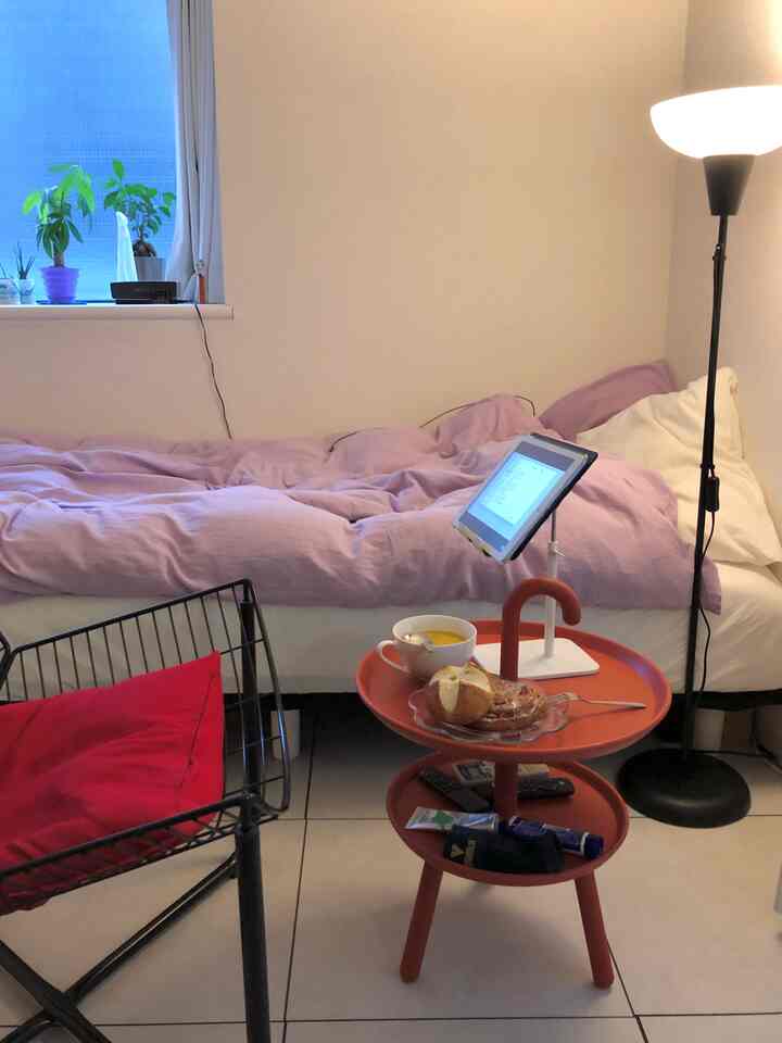 Cozy studio bedroom with white walls and purple bedding, featuring an orange tray table and floor lamp