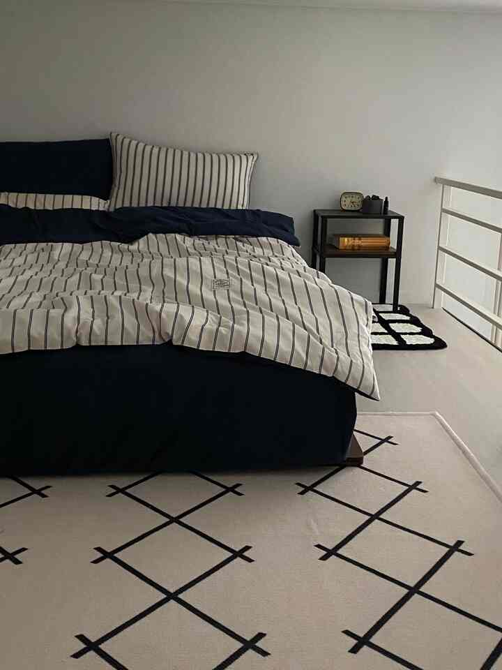 白い壁と床に黒とネイビーの寝具が調和するモダンな寝室、パターンラグとナイトテーブルが配置されたシンプルな空間