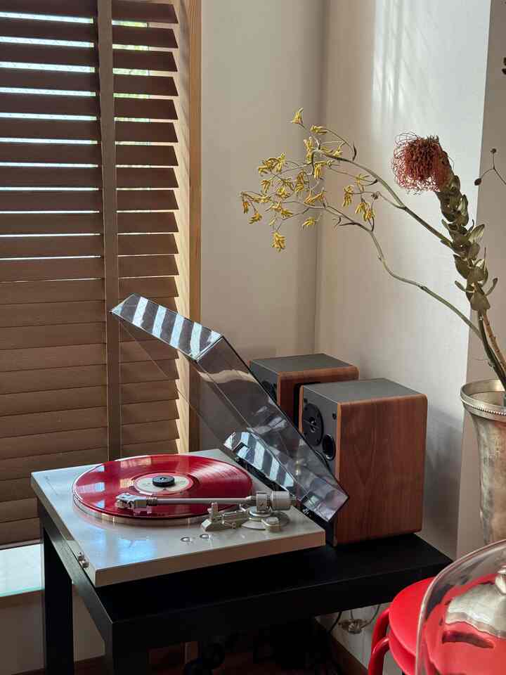 木目調のブラインドとサイドテーブルの上に赤いレコードがあるターンテーブルとスピーカーが配置された一人暮らしの1LDKの落ち着いた空間