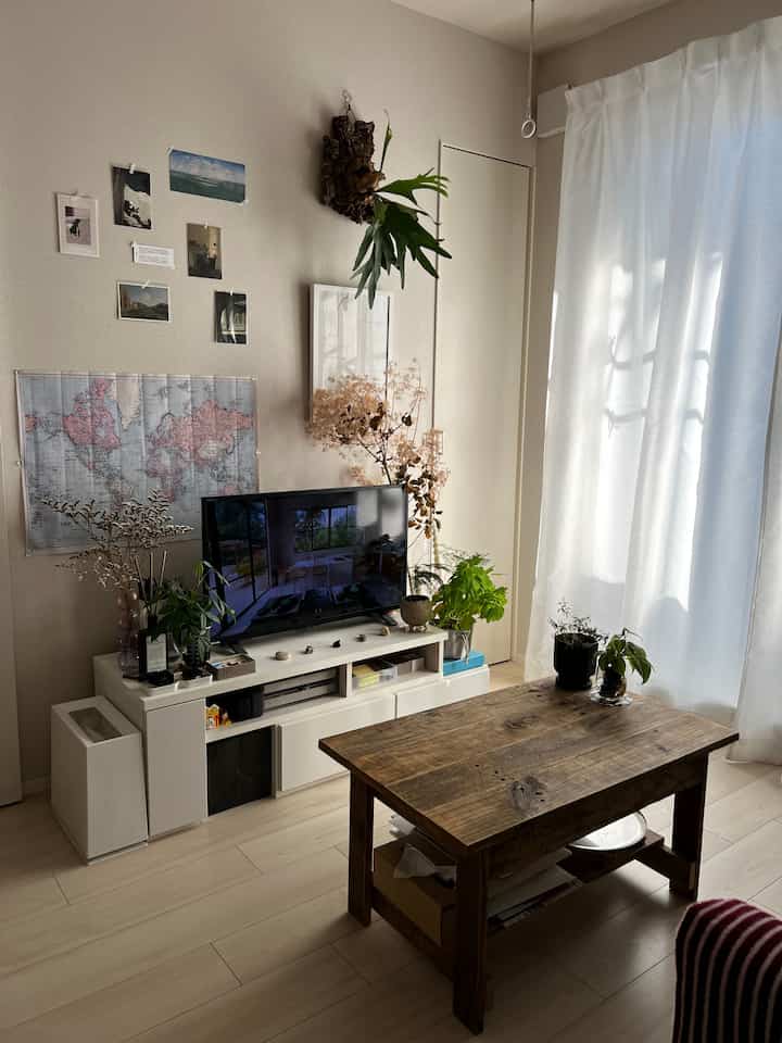 白い壁と明るい木目調のテーブルがあるナチュラルなリビング、植物とポスターが配置されて落ち着いた雰囲気のインテリア。