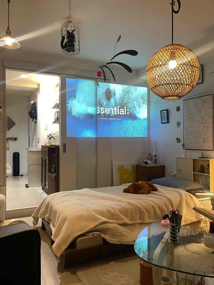 暖かいベージュとウッドトーンの一人暮らしリビング、ベッドとペンダントライト、プロジェクターが配置された居心地の良い空間