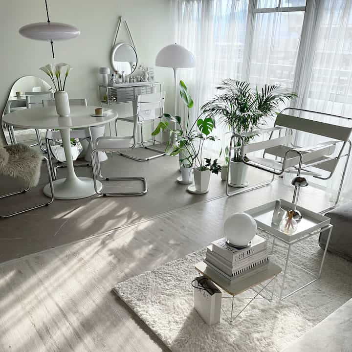 ホワイトとナチュラルトーンのリビング、ダイニングテーブルと椅子、多数の観葉植物が明るいカーテンの前に配置された雰囲気のあるインテリア